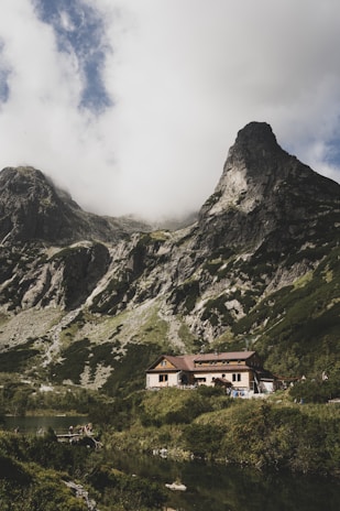 A rustic mountain lodge nestled among lush vegetation with towering rocky mountains in the background. The sky is partly cloudy, casting soft shadows over the landscape. A small body of water lies in the foreground, reflecting the serene environment.