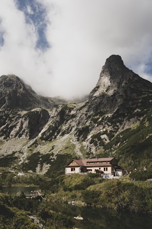 A rustic mountain lodge nestled among lush vegetation with towering rocky mountains in the background. The sky is partly cloudy, casting soft shadows over the landscape. A small body of water lies in the foreground, reflecting the serene environment.