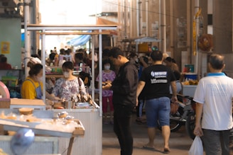 A bustling street market scene with several people interacting around food stalls. The central focus is on a vendor interacting with a customer while others are walking or browsing. The atmosphere is lively with a mix of bright colors from clothing and market wares. Some people are wearing masks, suggesting a recent or ongoing health concern.