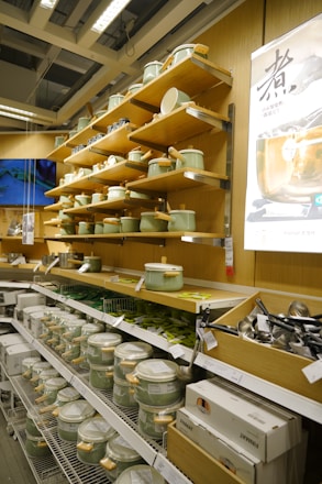 A well-organized retail display featuring shelves of cookware, including pots and pans with light green lids and wooden handles. The setup is in a clean, brightly lit store, with kitchen utensils, boxes, and promotional posters also visible.