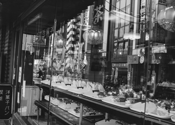 A black and white image of a bakery shop window displaying various baked goods on trays. The window reflects the bustling street outside, with visible signage and buildings. Traditional design elements suggest an older or culturally rich setting.