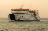 A large ferry named KMP Aceh Hebat 2 is sailing across calm waters under a clear sky. It has a white body with a red top and distinctive blue and yellow stripes. The vessel features multiple decks with visible railings and appears to be carrying passengers and vehicles.