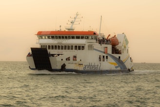A large ferry named KMP Aceh Hebat 2 is sailing across calm waters under a clear sky. It has a white body with a red top and distinctive blue and yellow stripes. The vessel features multiple decks with visible railings and appears to be carrying passengers and vehicles.
