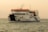 A large ferry named KMP Aceh Hebat 2 is sailing across calm waters under a clear sky. It has a white body with a red top and distinctive blue and yellow stripes. The vessel features multiple decks with visible railings and appears to be carrying passengers and vehicles.