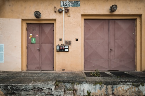 A weathered exterior wall of a building with two large, industrial metal doors, one smaller and one larger. Above the doors, there are two circular light fixtures. Between the doors, there is an electrical panel with several switches and a pipe running vertically. The wall is painted in a worn peach color, and there is a faded sign with the letters 'TS' followed by numbers. The ground in front of the building is concrete with some vegetation encroaching.