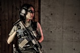A person wearing tactical gear, including headphones and protective glasses, holds a firearm while standing against a wooden wall adjacent to a concrete wall.