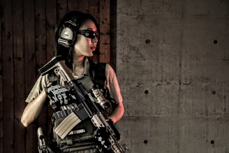 A person wearing tactical gear, including headphones and protective glasses, holds a firearm while standing against a wooden wall adjacent to a concrete wall.