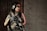 A person wearing tactical gear, including headphones and protective glasses, holds a firearm while standing against a wooden wall adjacent to a concrete wall.