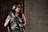 A person wearing tactical gear, including headphones and protective glasses, holds a firearm while standing against a wooden wall adjacent to a concrete wall.
