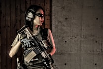 A person wearing tactical gear, including headphones and protective glasses, holds a firearm while standing against a wooden wall adjacent to a concrete wall.