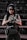 A person wearing tactical gear, including a helmet, sunglasses, and a headset, standing on stairs. They are holding a firearm and have a tactical vest with pouches. The setting appears to be outdoors with a dark, weathered backdrop.