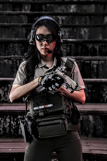 A person wearing tactical gear, including a helmet, sunglasses, and a headset, standing on stairs. They are holding a firearm and have a tactical vest with pouches. The setting appears to be outdoors with a dark, weathered backdrop.