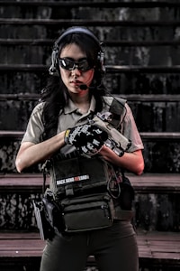 A person wearing tactical gear, including a helmet, sunglasses, and a headset, standing on stairs. They are holding a firearm and have a tactical vest with pouches. The setting appears to be outdoors with a dark, weathered backdrop.