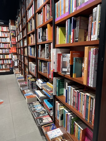 A well-organized bookstore aisle lined with wooden shelves packed with various books. Many of the books are neatly arranged standing upright, while others are stacked horizontally on the lower shelves. The books cover a range of topics and are assorted in different colors, creating a vibrant and inviting atmosphere. Shadows are cast by the shelving, adding depth to the scene.