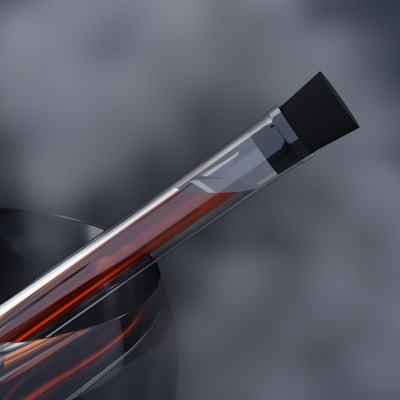 A transparent test tube partially filled with a dark red liquid is angled diagonally against a blurred, grayish background. The test tube is capped with a black stopper and is placed inside a swirling, semi-transparent material, possibly a holder or container.