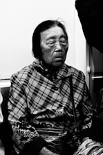 An elderly person with closed eyes sits in what appears to be a public transportation setting. The individual wears a patterned jacket, possibly plaid, and holds a textured purse. The lighting is low, giving a reflective, calm atmosphere, and the image is in black and white.