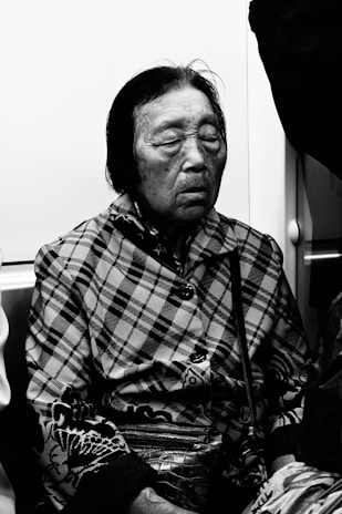 An elderly person with closed eyes sits in what appears to be a public transportation setting. The individual wears a patterned jacket, possibly plaid, and holds a textured purse. The lighting is low, giving a reflective, calm atmosphere, and the image is in black and white.