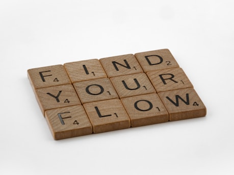 Wooden Scrabble tiles arranged in three rows to spell out the phrase 'FIND YOUR FLOW'. Each tile has a letter with a corresponding numerical value in the bottom right corner.