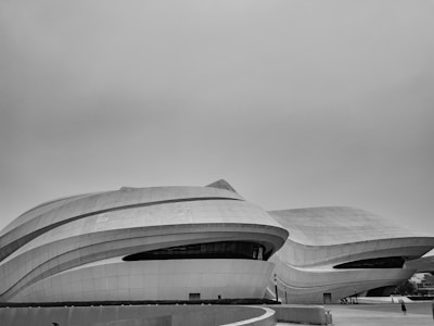A futuristic, modern architectural structure with smooth, curved surfaces and large window sections. The buildings appear to be made of metallic or concrete materials, with a sleek, spaceship-like design. The sky is overcast, adding a monochromatic feel to the scene.