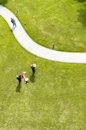 Aerial view of a well-maintained grassy area with a curved pathway. Two people wearing hats and green uniforms are tending to the lawn, with gardening tools and a red bucket nearby. Another person is walking along the path carrying a backpack.