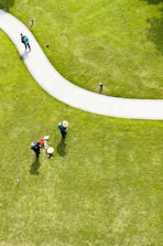 Aerial view of a well-maintained grassy area with a curved pathway. Two people wearing hats and green uniforms are tending to the lawn, with gardening tools and a red bucket nearby. Another person is walking along the path carrying a backpack.