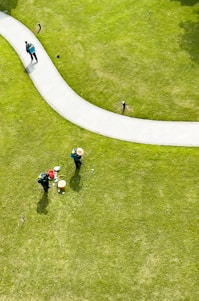 Aerial view of a well-maintained grassy area with a curved pathway. Two people wearing hats and green uniforms are tending to the lawn, with gardening tools and a red bucket nearby. Another person is walking along the path carrying a backpack.