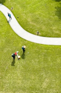 Aerial view of a well-maintained grassy area with a curved pathway. Two people wearing hats and green uniforms are tending to the lawn, with gardening tools and a red bucket nearby. Another person is walking along the path carrying a backpack.