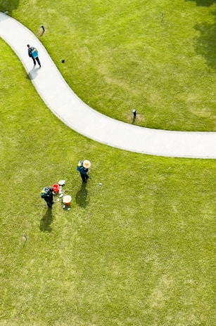Aerial view of a well-maintained grassy area with a curved pathway. Two people wearing hats and green uniforms are tending to the lawn, with gardening tools and a red bucket nearby. Another person is walking along the path carrying a backpack.