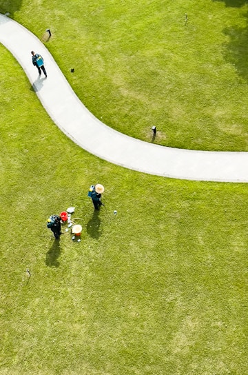 Aerial view of a well-maintained grassy area with a curved pathway. Two people wearing hats and green uniforms are tending to the lawn, with gardening tools and a red bucket nearby. Another person is walking along the path carrying a backpack.