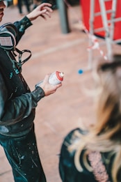 A person holding a spray paint can appears to be in the midst of a painting project. They are wearing a respirator mask around their neck and have paint splatters on their clothing. In the background, there is another person with blonde hair partially visible, a red structure, and a cordon tape.