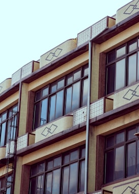 The image showcases a segment of a building fa&ccedil;ade featuring large rectangular glass windows framed with dark materials. The structure includes decorative geometric patterns on light-colored surfaces between the windows. The architectural style suggests a mix of modern and traditional elements, possibly indicating an office or residential building. There are metal fixtures, likely part of a drainage system, running down along the outer wall.