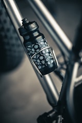 A close-up of a black water bottle with white symbols attached to a metallic bicycle frame. The bottle is secured in a holder, with part of the bike’s tire visible in the background.