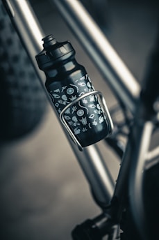 A close-up of a black water bottle with white symbols attached to a metallic bicycle frame. The bottle is secured in a holder, with part of the bike’s tire visible in the background.