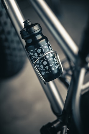 A close-up of a black water bottle with white symbols attached to a metallic bicycle frame. The bottle is secured in a holder, with part of the bike&rsquo;s tire visible in the background.