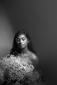 A monochromatic portrait captures a serene woman with long hair holding a bouquet of delicate flowers. Her eyes are closed, and the gentle play of light and shadow creates a tranquil atmosphere.