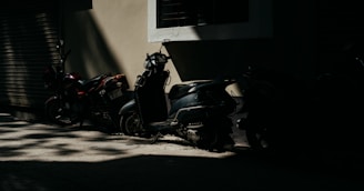 Two scooters and a motorcycle are parked in a dimly lit alley against a beige wall with a partially open roller shutter. The light creates a pattern of shadows on the ground and highlights portions of the vehicles, emphasizing the contrast between light and dark.
