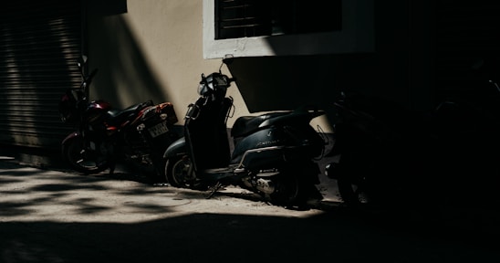 Two scooters and a motorcycle are parked in a dimly lit alley against a beige wall with a partially open roller shutter. The light creates a pattern of shadows on the ground and highlights portions of the vehicles, emphasizing the contrast between light and dark.