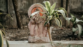 A decorative statue is placed in a garden setting, surrounded by small plants. The statue features detailed carvings and appears to be made of a stone material, resting on a concrete ledge. The surrounding area is filled with soil and plant life, contributing to a serene, natural environment.