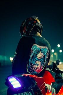 A person wearing a helmet sits on a red motorcycle at night. The helmet and jacket have vibrant designs, featuring illustrations on the back. The scene is illuminated by streetlights, casting a moody and atmospheric glow.