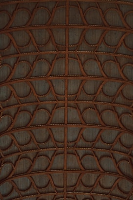A complex arrangement of interwoven wooden arches and beams, forming a symmetrical pattern. The wood has a natural brown tone, and the background displays a weathered, pale blue surface.