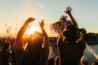 A crowd enjoys a live outdoor concert during sunset. The sun casts a warm glow over the audience with raised hands silhouetted against the sky. A stage with a performance is visible in the background, surrounded by tall palm trees.