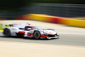 A racing car speeds down a track, exhibiting motion blur that emphasizes its high speed. The vehicle is designed aerodynamically, with sponsor logos visible on its body. The track is bordered by colorful barriers in red, yellow, and orange.