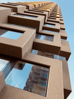 A modern architectural structure featuring a repetitive geometric pattern of cubic elements projecting outward. The building is composed of textured, light brown material with large reflective glass windows, contrasting with the clear blue sky in the background.