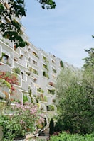 A building with a unique architectural design, featuring numerous terraces adorned with lush plants and flowers. The structure appears to be an apartment complex, with each unit having its own private green space. The variety of plants includes trees, shrubs, and flowering plants, adding vibrant greenery and colors to the facade. The setting is serene, framed by additional trees and a clear blue sky.