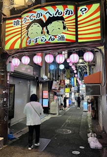 A vibrant, illuminated street scene features a large, colorful sign with cartoon faces and Japanese text, set against a background of red and yellow radial stripes. Below the sign, several pink lanterns are strung across the alley, which is lined with various shops and illuminated signs. People are walking down the alley, creating a lively atmosphere.