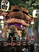 A group of people wearing traditional clothing perform on a decorated wooden float adorned with intricate carvings. The float is illuminated by lanterns that feature bold Japanese characters. The setting appears to be outdoors, under a tree, possibly during a festival or cultural event.