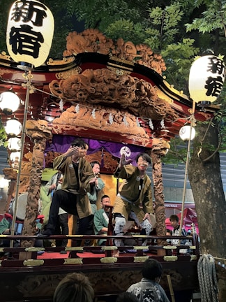 A group of people wearing traditional clothing perform on a decorated wooden float adorned with intricate carvings. The float is illuminated by lanterns that feature bold Japanese characters. The setting appears to be outdoors, under a tree, possibly during a festival or cultural event.