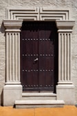 A robust wooden door framed by ornate stone pillars built into a beige stone facade. The door is dark brown and features a series of evenly spaced round metal studs and a central metallic handle.