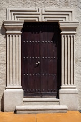 A robust wooden door framed by ornate stone pillars built into a beige stone facade. The door is dark brown and features a series of evenly spaced round metal studs and a central metallic handle.