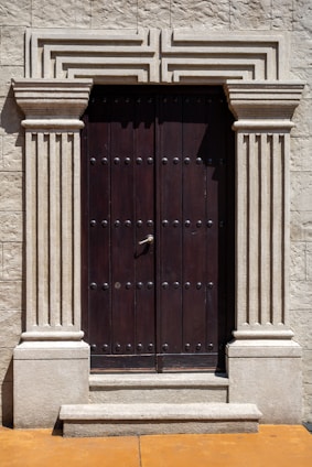 A robust wooden door framed by ornate stone pillars built into a beige stone facade. The door is dark brown and features a series of evenly spaced round metal studs and a central metallic handle.